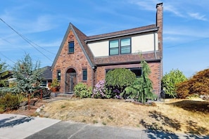 A two-story brick house with a front entrance, a visible chimney, and a well-maintained garden with various shrubs and a lawn. - 2 BR Home in the heart of Beacon Hill (Seattle)