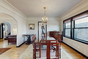 A spacious dining area with a long table and chairs, a chandelier, and large windows offering a view. - 2 BR Home in the heart of Beacon Hill (Seattle)