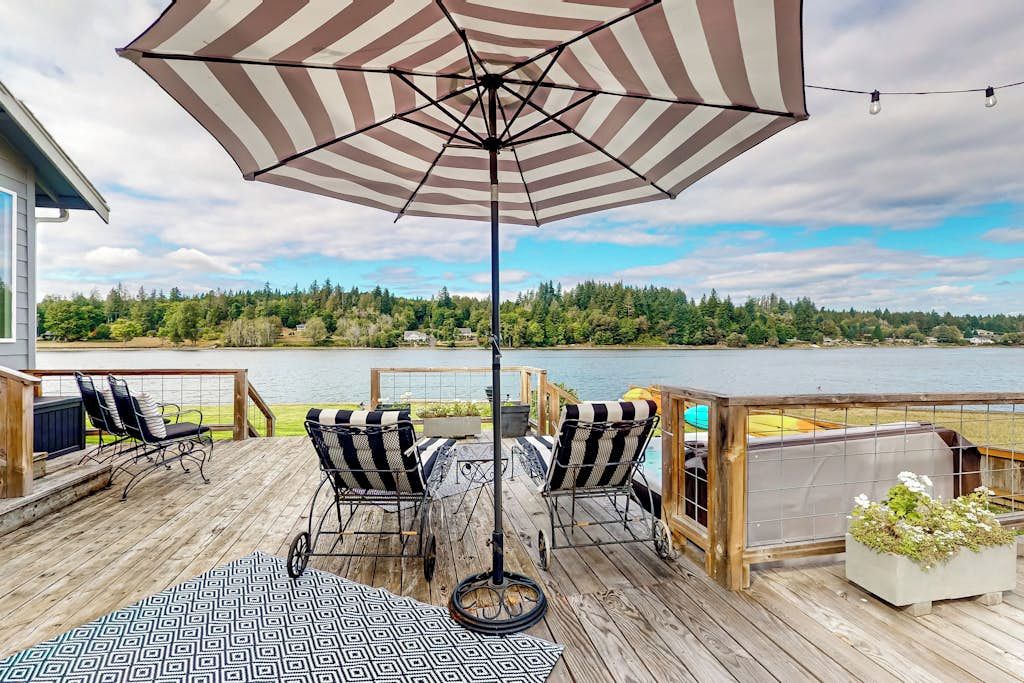 Een houten terras met een grote gestreepte parasol, zitplaatsen buiten en uitzicht op een meer.