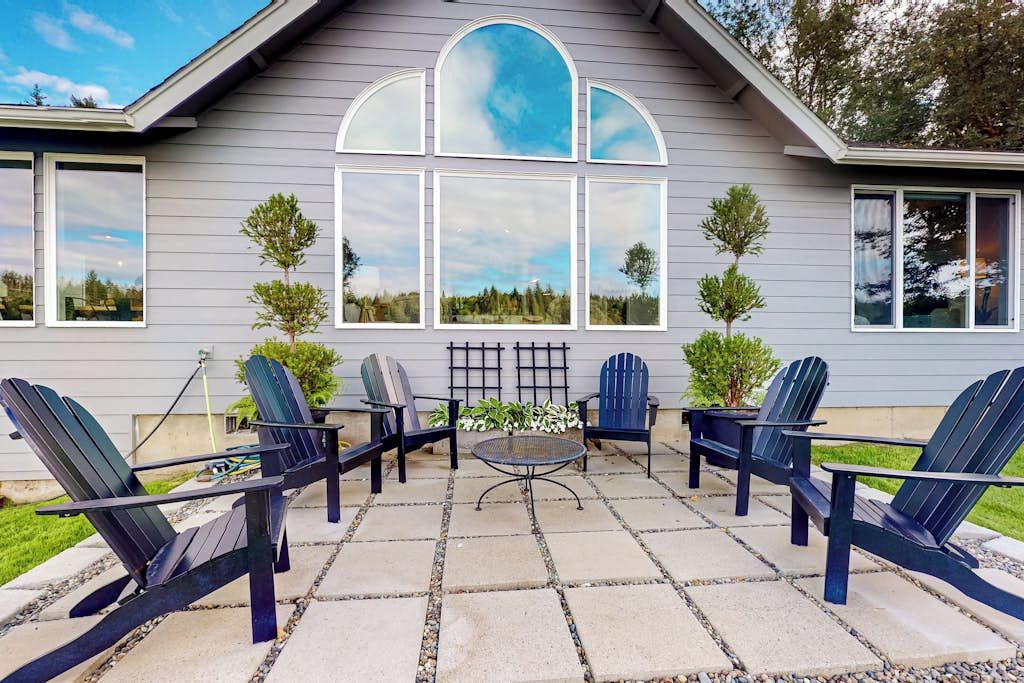 A patio with Adirondack chairs and a table, a large window with a view, and a small tree.