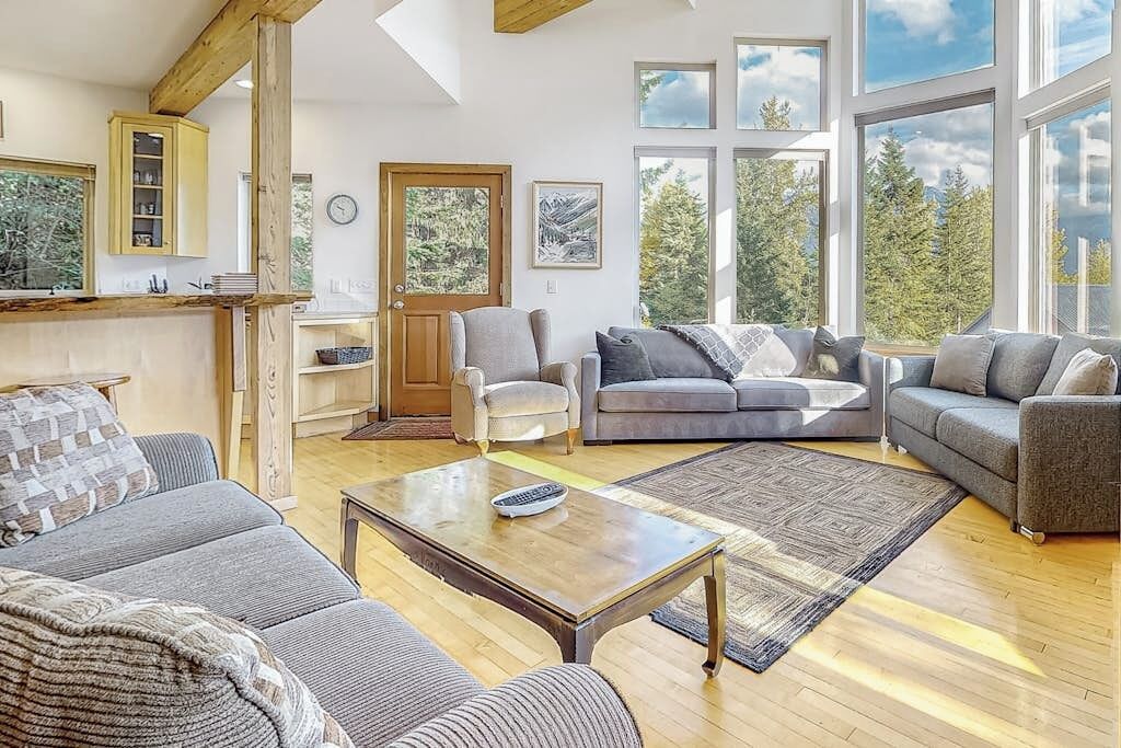 Een ruime woonkamer met een houten salontafel, grijze banken en uitzicht op bomen door grote ramen.