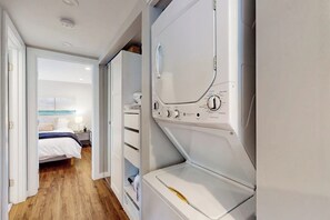 Ein Waschraum mit Waschmaschine und Trockner, ein Bett mit weißer Bettwäsche und Blick in ein Schlafzimmer durch eine geöffnete Tür.