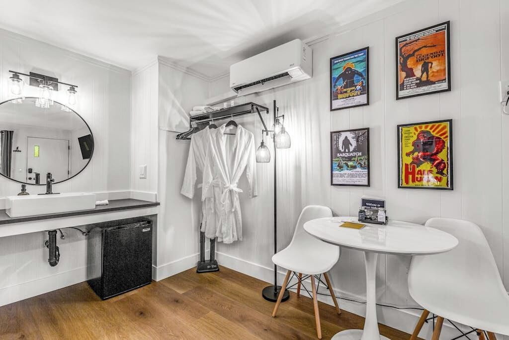 A modern bathroom with a large mirror, white sink, and a black refrigerator. There are two white chairs and a small table with a sign.