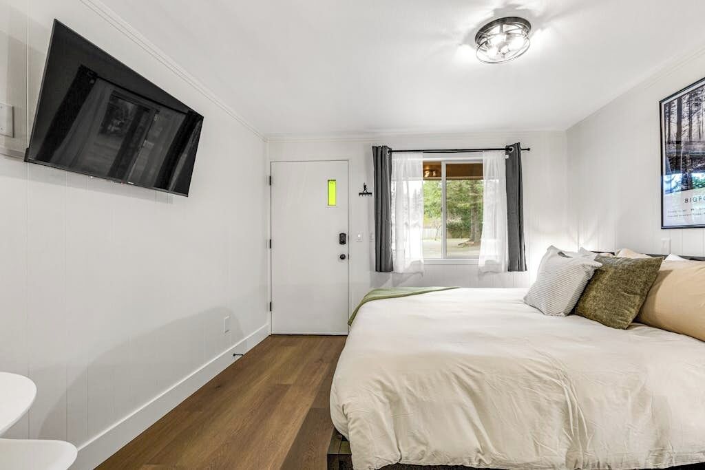 Quarto com cama, televisão na parede, porta e janela com cortinas.