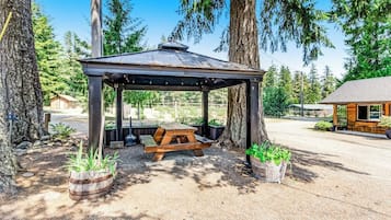 A covered picnic area with a bench and picnic table, surrounded by trees and a building in the background.