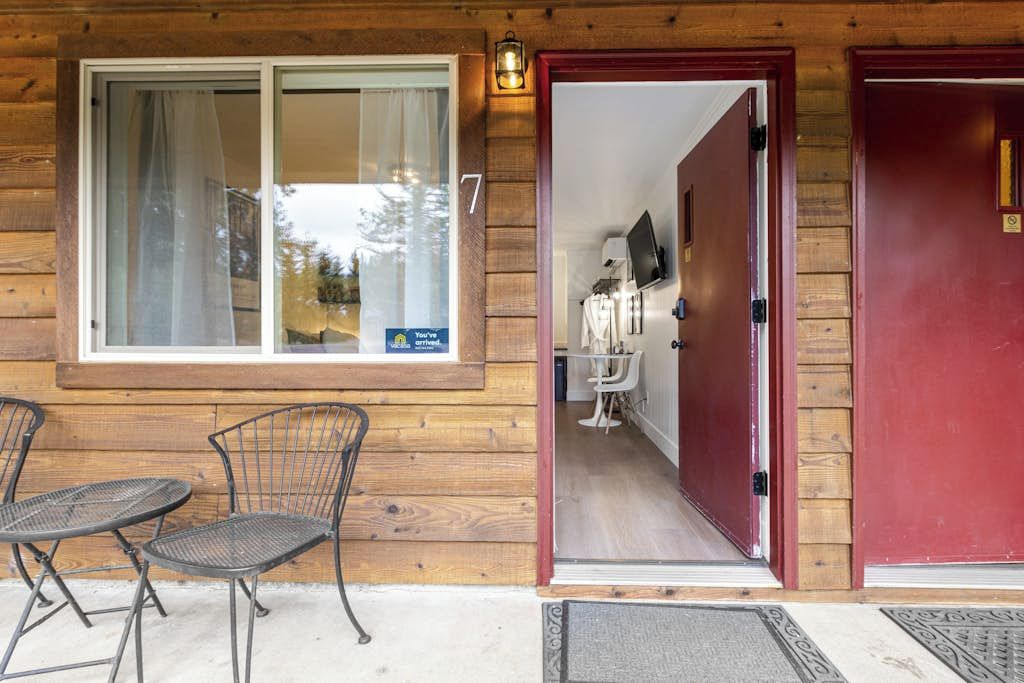 Fachada de madera con el número 7, puerta de cristal que da a una habitación y una pequeña zona de estar al aire libre con sillas de metal.