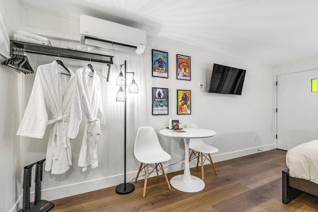 Habitación de hotel con cama, una mesa redonda con dos sillas, una lámpara de pie, televisor montado en la pared y un perchero con batas blancas.