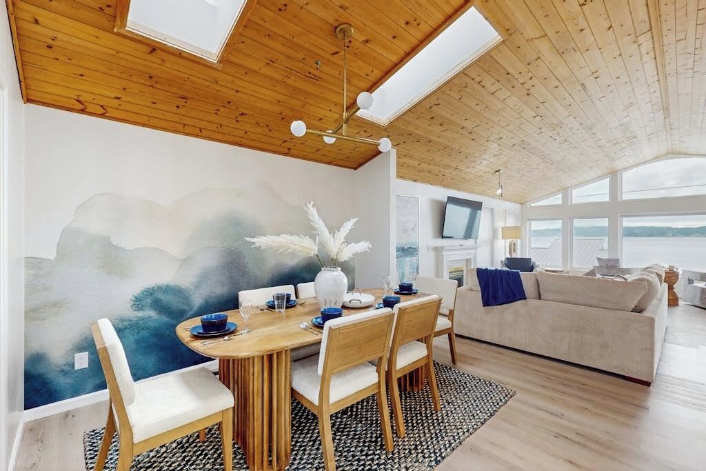 A modern living room with a wooden ceiling, a dining table with chairs, a sofa, and a large window offering a view.