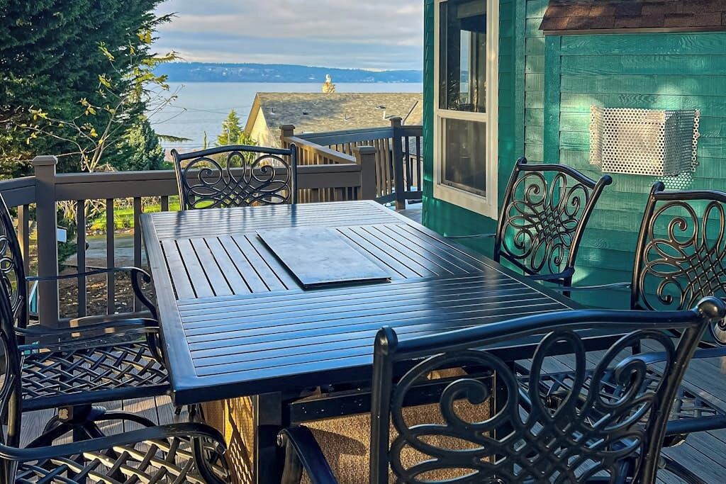 Eine Holzterrasse mit Tisch und Stühlen, mit Blick auf ein Gewässer und entfernte Gebäude.