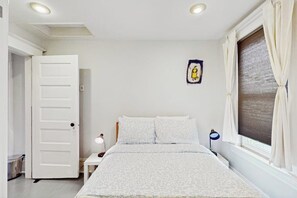 Ein Schlafzimmer mit Bett, Nachttisch, Lampe, einem Fenster mit Vorhängen und einer Tür.
