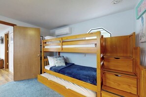 A bunk bed with a wooden frame, a blue blanket, and a pillow with 'Beach House' text. There is a wooden cabinet to the right and a door to the left.