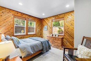 A bedroom with a herringbone-patterned wooden wall, a bed with a grey bedspread, a wooden dresser, and two windows with potted plants. - Lakefront views with hot tub, firepit, & deck (Sedro-Woolley)