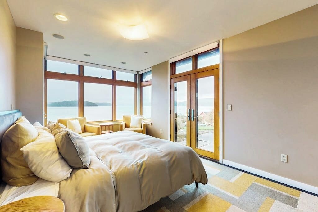 A bedroom with a large bed, a seating area with a table, and a view through a glass door.