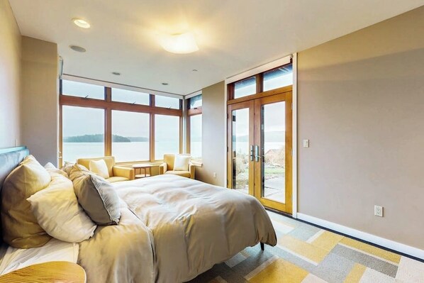 A bedroom with a large bed, a seating area with a table, and a view through a glass door.