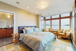 A bedroom with a large bed, a sitting area with a sofa and armchairs, a small table, and a view of the outdoors through large windows.