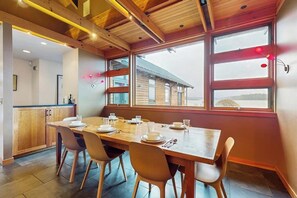 A dining area with a wooden table and chairs, a kitchen in the background, and large windows with a view of a building outside.