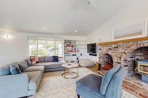 A modern living room with a sectional sofa, a round coffee table, a brick fireplace, and a bookshelf. - 2BR Waterfront Home with Private Beach Access (Belfair)