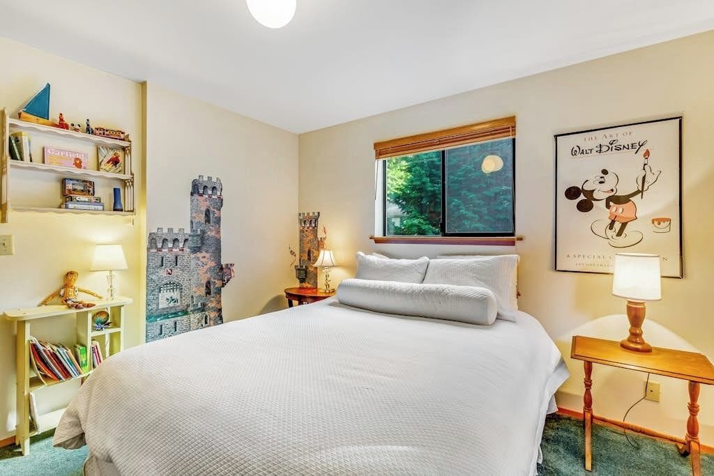 A hotel room with a large bed, a bookshelf, a castle mural, a window with a view, and a poster of Walt Disney.