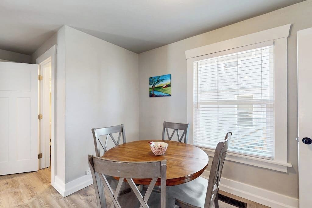 A dining area with a round wooden table, four chairs, a bowl of fruit, and a window with blinds.