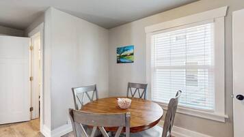 A dining area with a round wooden table, four chairs, a bowl of fruit, and a window with blinds.