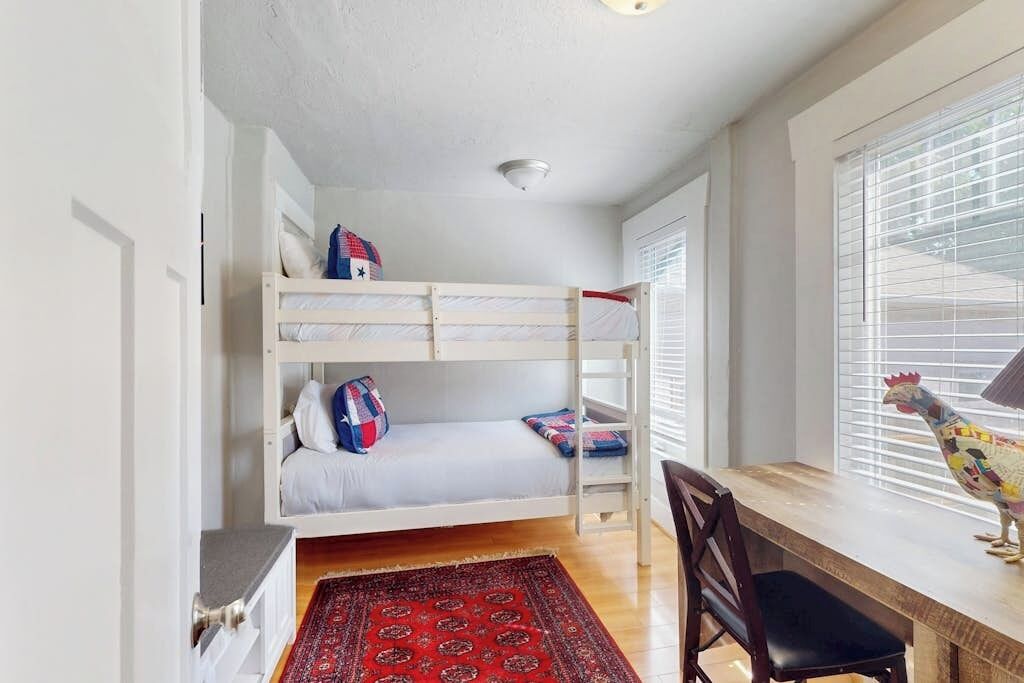A bunk bed with pillows, a desk with a chair, a rooster decoration, and a window with blinds.