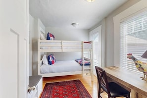 A bunk bed with pillows, a desk with a chair, a rooster decoration, and a window with blinds.