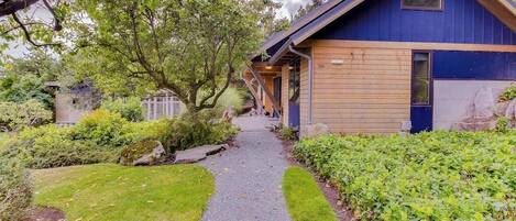 A wooden cabin with a gravel path leading to the entrance, surrounded by lush greenery and trees.