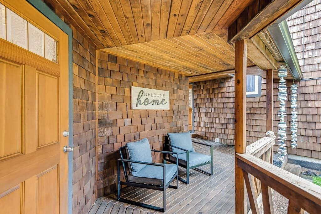 A wooden deck with two chairs, a wooden door, and a 'Welcome home' sign on a brick wall.
