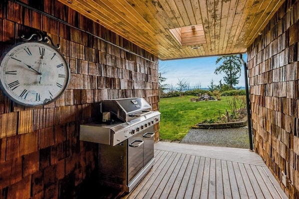 A wooden deck with a barbecue grill and a wall-mounted clock. - Idyllic woodsy 6BR with firepit & two kitchens (Camano Island)
