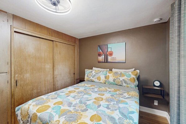Un dormitorio con cama, armario de madera, mesita de noche con un reloj y una obra de arte en la pared.