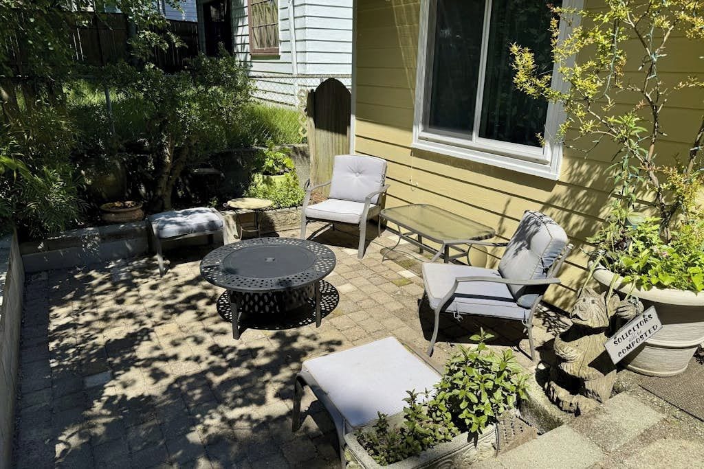 An outdoor patio with a round table, two chairs, and a small table. There are potted plants and a statue.
