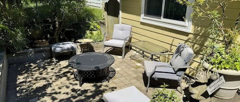 Une terrasse extérieure avec une table ronde, deux chaises et une petite table d’appoint. On y trouve également des plantes en pot et une statue.