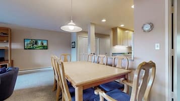 A dining area with a wooden table and chairs, a television on the left, and a kitchen in the background.
