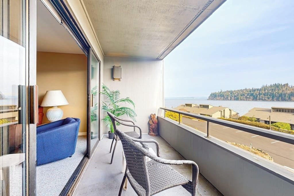 Un balcon donnant sur une étendue d’eau et une côte lointaine, un canapé bleu et deux fauteuils en osier.