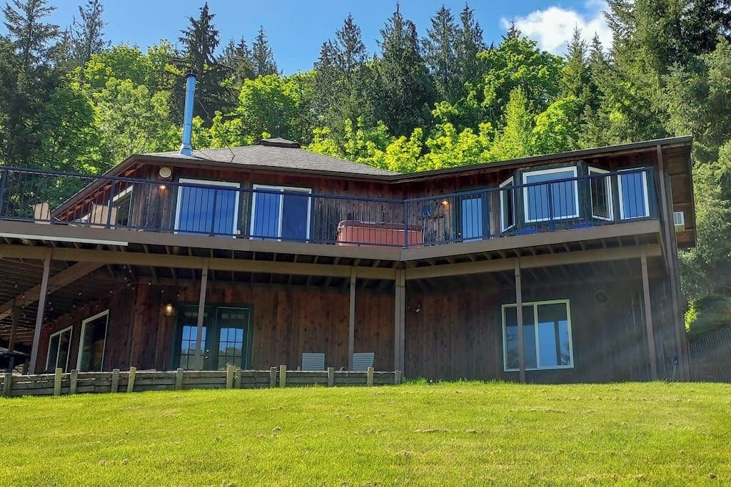 Una cabaña de madera de dos pisos con balcón, rodeada por un patio cubierto de césped y un bosque denso.