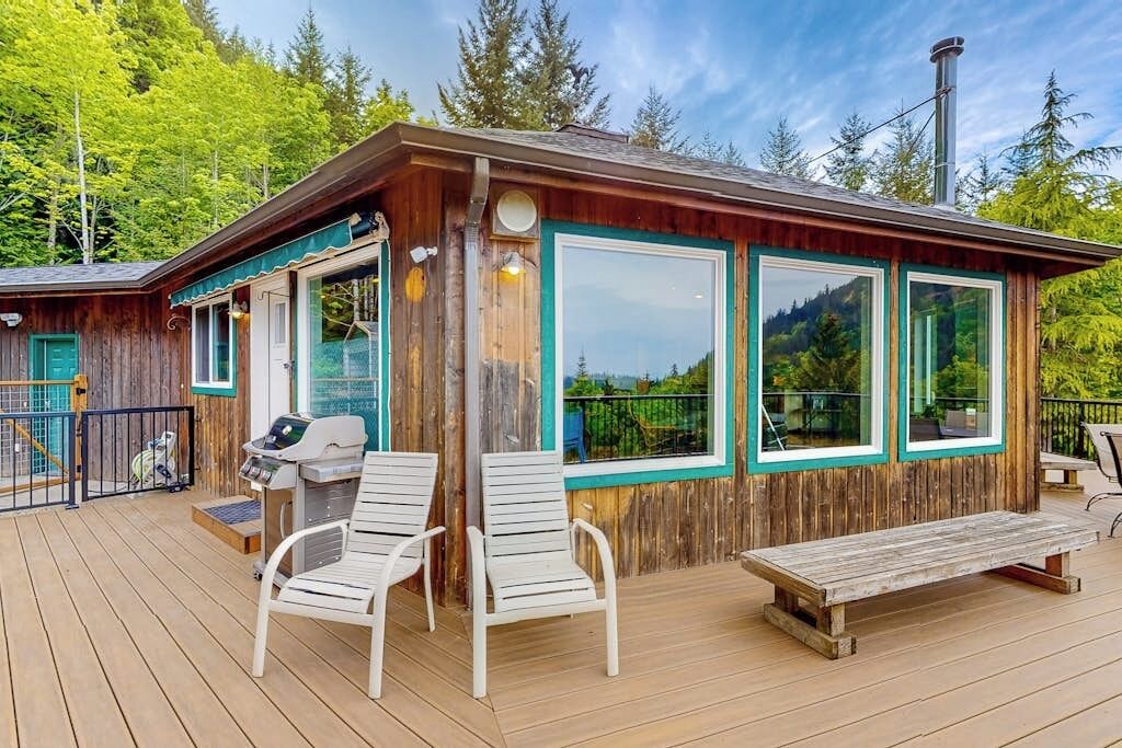 Une cabane en bois avec une terrasse, deux chaises blanches, un banc et un barbecue.