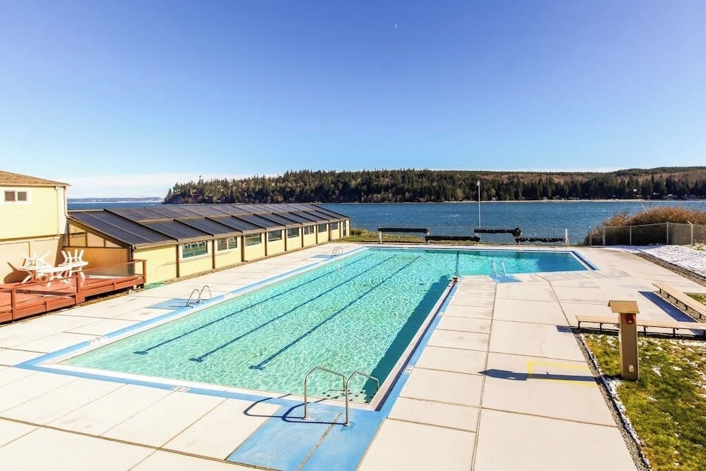 A swimming pool with a view of a lake and forest, a covered area with chairs, and a building with a sloped roof.