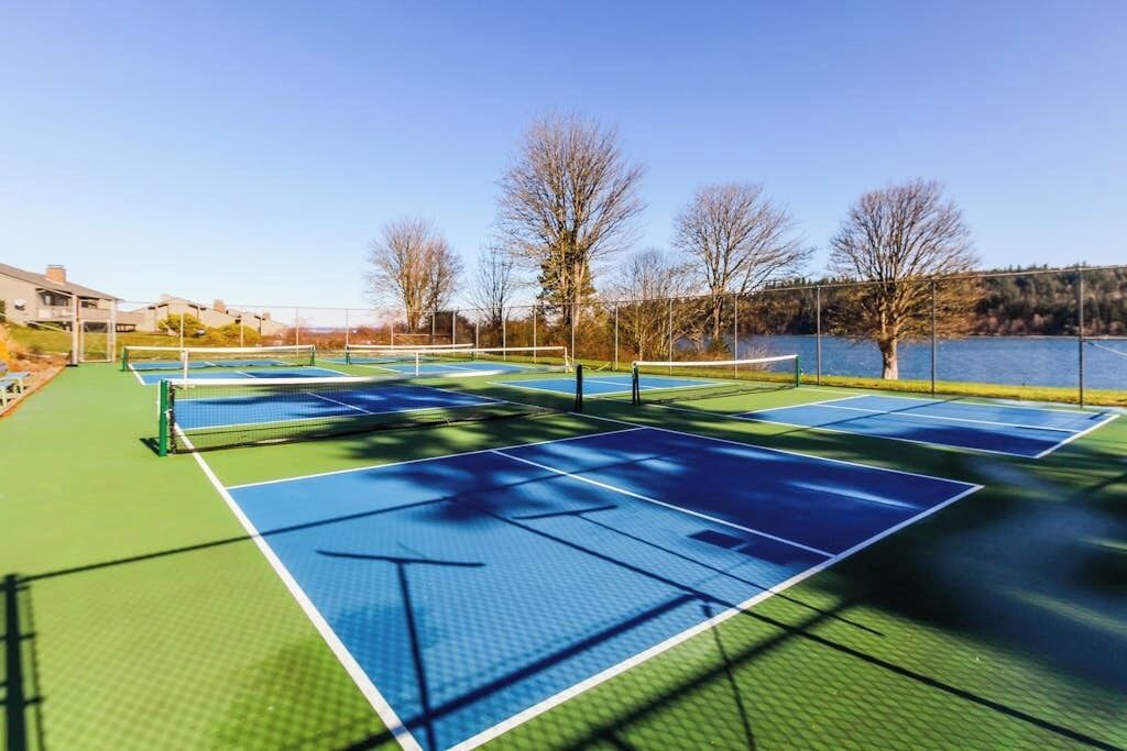 Una cancha de tenis exterior con varias canchas, rodeada de árboles y un cuerpo de agua.