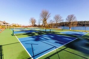 An outdoor tennis court with multiple courts, surrounded by trees and a body of water.