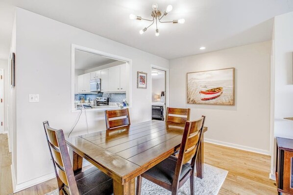A dining area with a wooden table and chairs, a kitchen in the background, and a framed artwork on the wall.