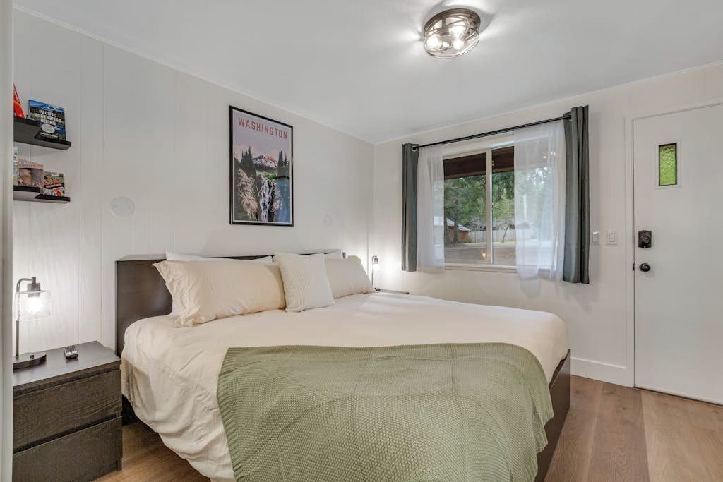 Quarto com cama, mesa de cabeceira, janela com cortinas, porta e prateleira com livros.