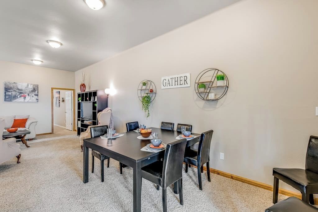 A dining area with a table set for four, surrounded by chairs, with a 'GATHER' sign on the wall and a bookshelf in the background.