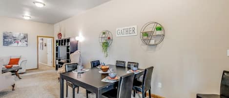Un coin repas avec une table dressée pour quatre personnes, entourée de chaises, et un panneau « RASSEMBLEMENT » accroché au mur, avec une bibliothèque en arrière-plan.
