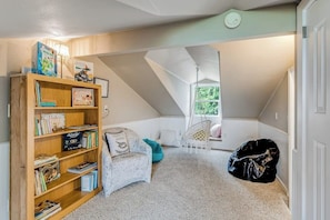 A cozy attic bedroom with a sloped ceiling, a bookshelf filled with books, a patterned armchair, and a window with a view. - Island 3BR with firepit & port & mountain views (Camano Island)