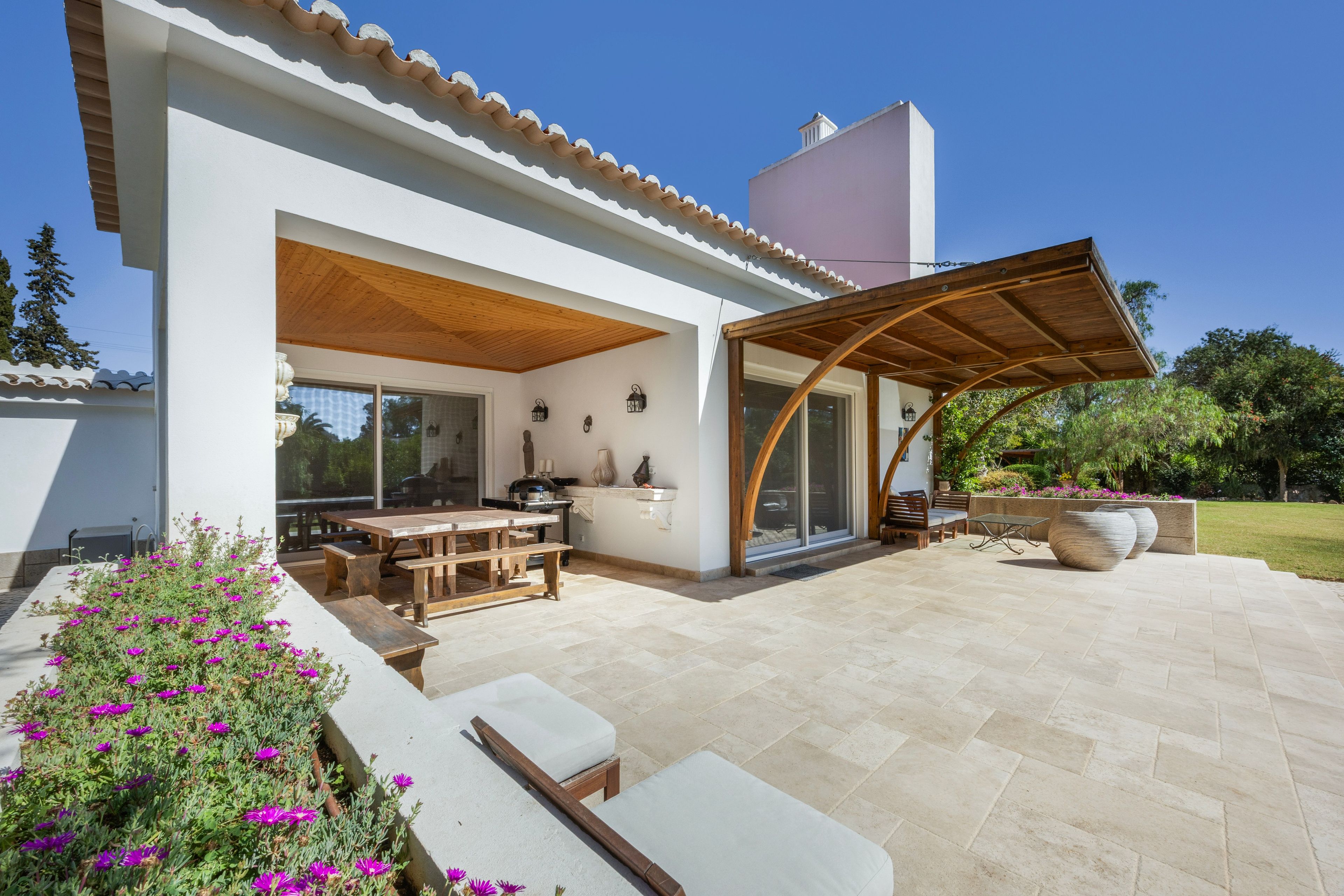 Un patio moderno al aire libre con una mesa y bancos de madera, una zona de estar cubierta y un jardín con flores moradas.