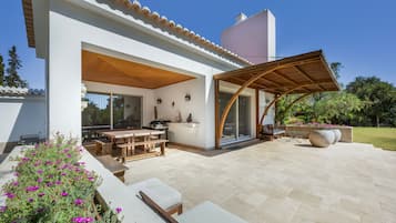 Un patio moderno al aire libre con una mesa y bancos de madera, una zona de estar cubierta y un jardín con flores moradas.