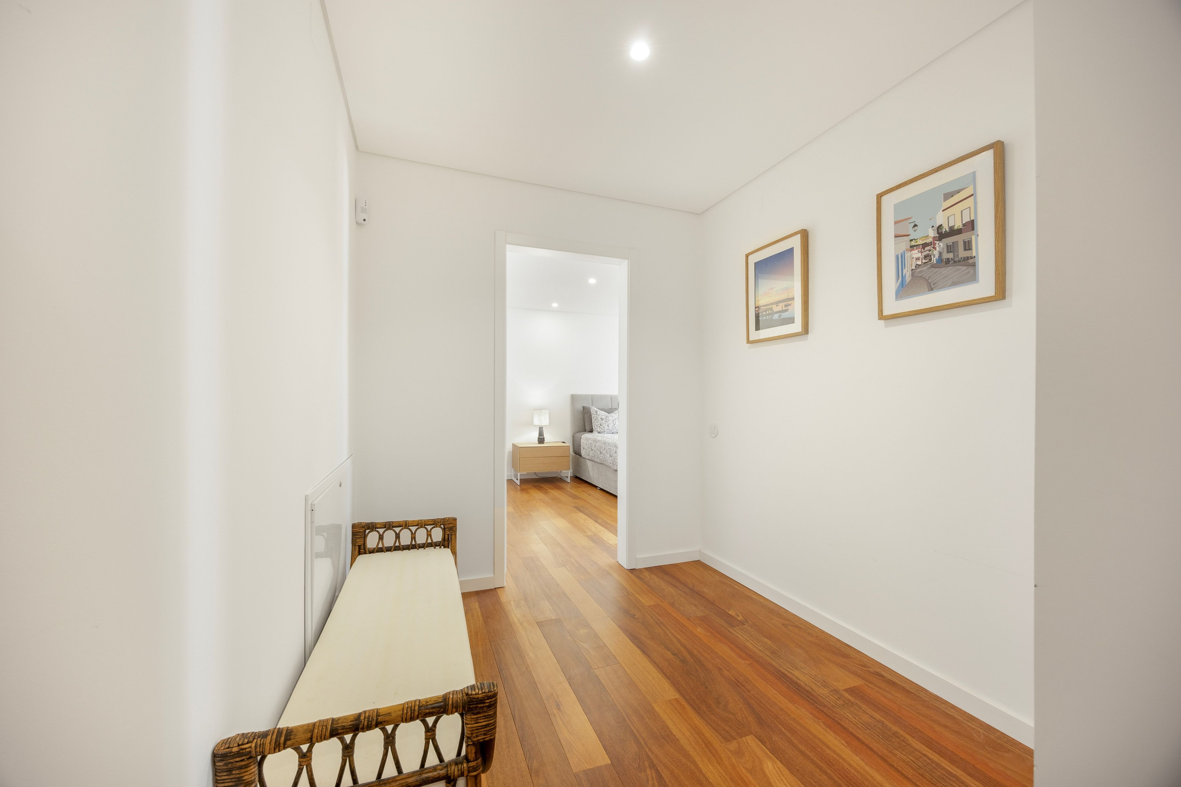 A hallway with a wooden floor, a bed with a white headboard, and two framed pictures on the wall.