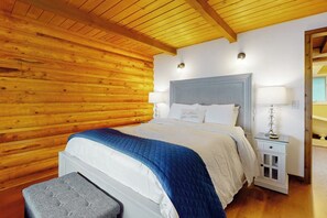 A bedroom with a wooden ceiling, a bed with a blue and white bedspread, a bedside table with a lamp, and a nightstand with a lamp.