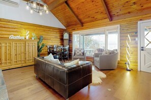 A log cabin interior with a leather sofa, a dining area, and a view through the windows.