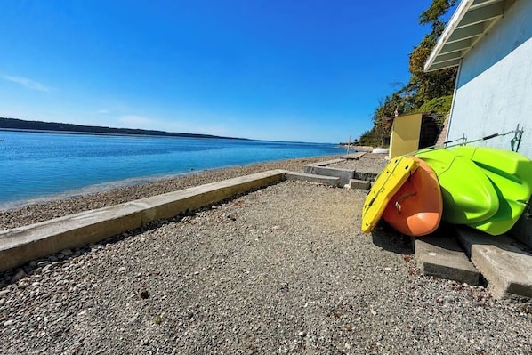 A pebbled beach with a concrete walkway, a building with a white exterior, and a calm body of water. - Sweeping 2BR Oceanfront - WoodStove - Balcony (Camano Island)